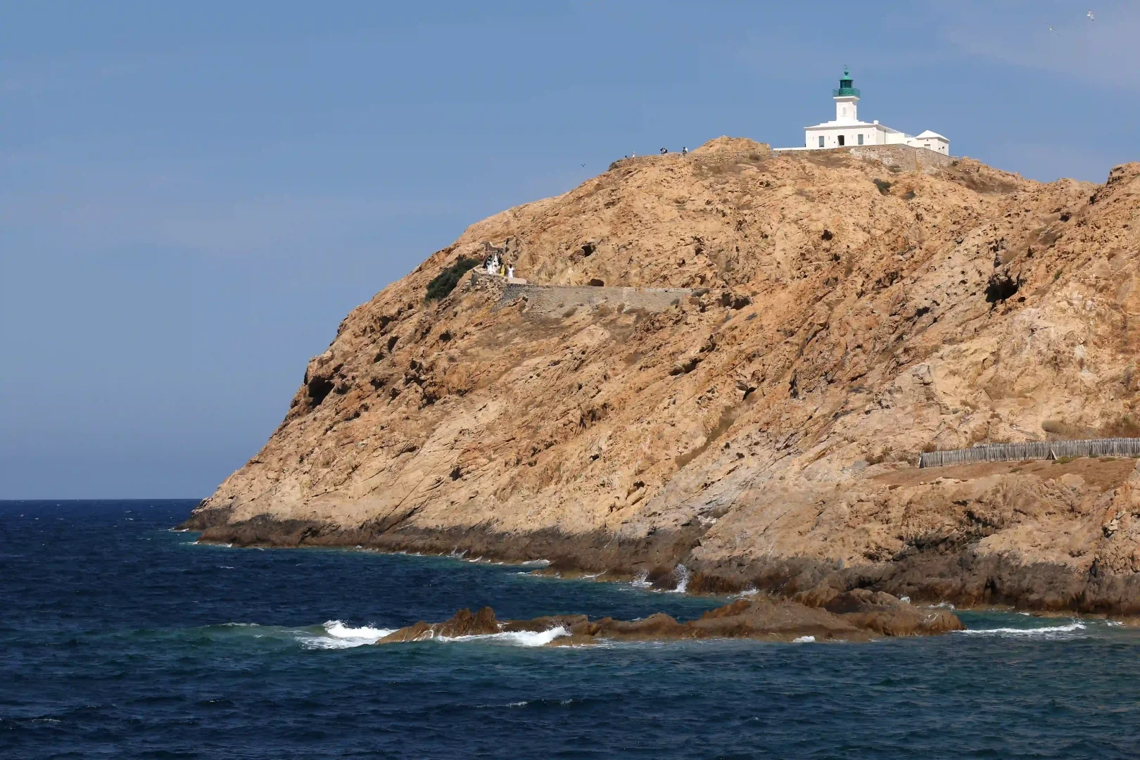 Korsika kıyılarında denize uzanan kayalık bir tepenin üzerinde yükselen, beyaz gövdeli ve yeşil kubbeli tarihi Phare de la Pietra deniz feneri, Jacquemus'un Akdeniz'in doğal güzelliğini ve yazın hafifliğini kutlayan Le Bonheur koleksiyonu için etkileyici bir fon oluşturuyor.