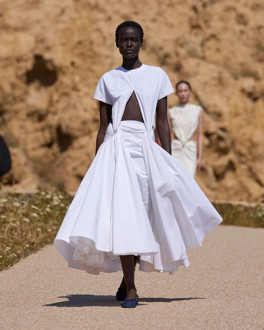 Koyu tenli bir model, Jacquemus'un Le Bonheur İlkbahar-Yaz 2027 koleksiyonundan karın dekolteli beyaz kısa kollu üstü ve beli sarmalayan, hafifliği vurgulayan hacimli, katmanlı açılan etekle, güneşli, doğal kayalık bir fon önünde kum rengi podyumda kendinden emin adımlarla ilerliyor.