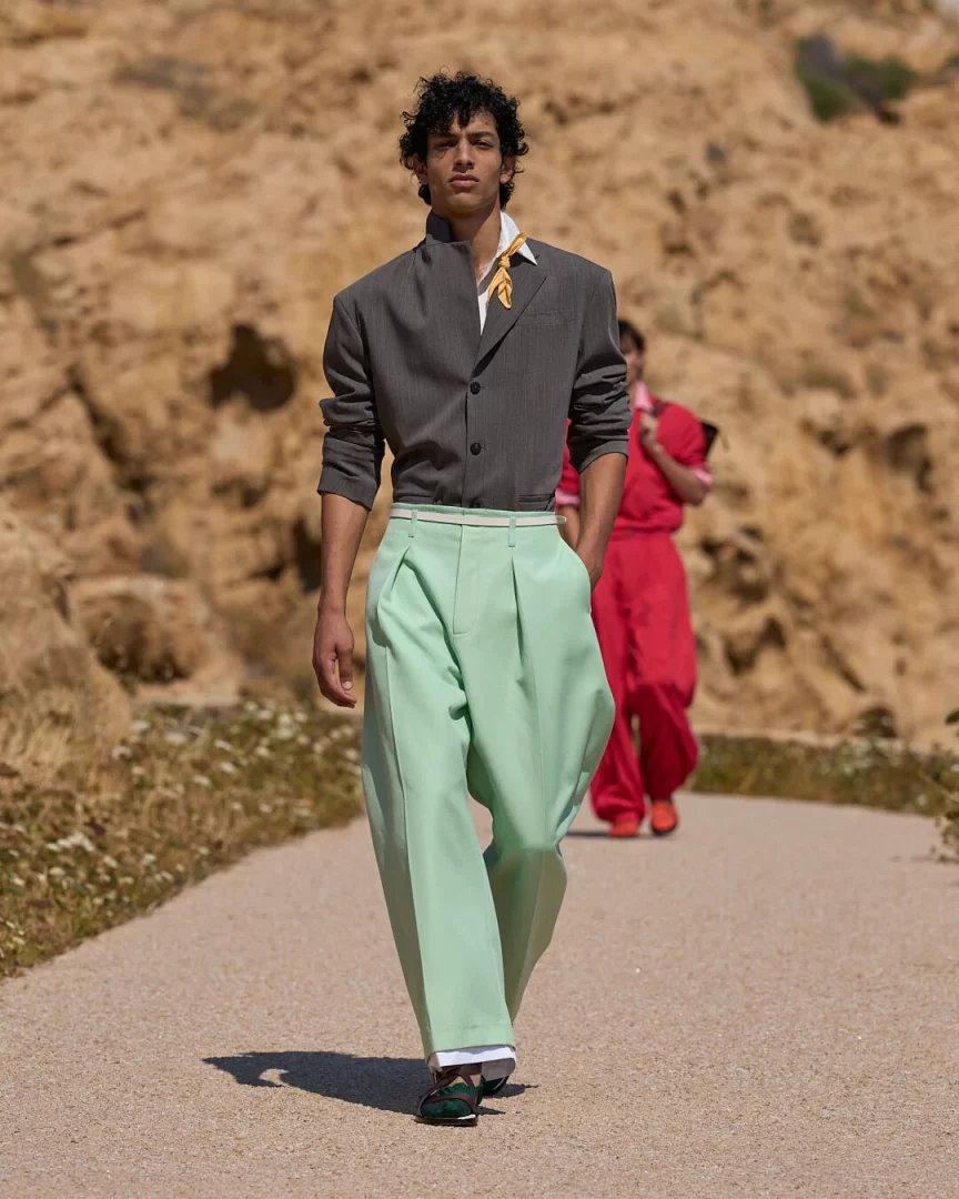 Kıvırcık saçlı bir erkek model, Jacquemus İlkbahar-Yaz 2027 Le Bonheur koleksiyonundan gri bir ceket, sarı eşarp detaylı beyaz gömlek ve nane yeşili bol pantolonla doğal, kayalık Akdeniz manzarasında bir defile podyumunda yürüyor; arkada bulanık bir şekilde başka bir model kırmızı giysiyle görülürken, hafiflik ve yaz ruhunu yansıtan canlı renkler öne çıkıyor.