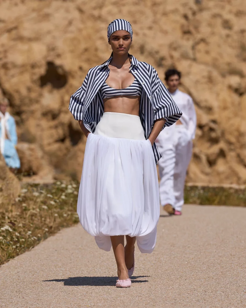 Jacquemus'un Akdeniz estetiğini yeniden tanımlayan Le Bonheur İlkbahar-Yaz 2027 koleksiyonundan bir görünüm sergileyen model, çizgili başörtüsü, bralet, bol gömlek ve heykelsi hacimli beyaz organza etekle doğal bir defile lokasyonunda zarafetle yürüyor.