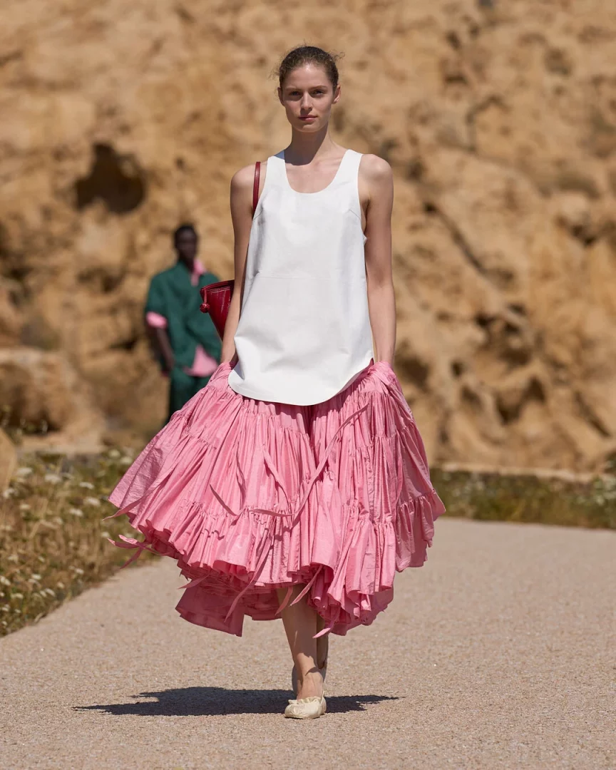 Jacquemus İlkbahar-Yaz 2027 defilesinden bir model, hafif beyaz atleti, rüzgarla hareket eden hacimli pembe fırfırlı eteği ve omzundaki küçük kırmızı çantasıyla doğal bir patikada Akdeniz'in hafifliğini yansıtan bir yürüyüş sergiliyor.