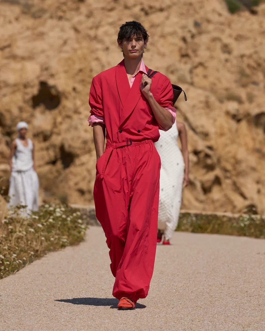 Jacquemus İlkbahar-Yaz 2027 Le Bonheur koleksiyonundan, Akdeniz'in doğal manzarasında hafiflik ve canlı renklerle sunulan bir görünüm; genç bir erkek model, açık pembe gömleği üzerine giydiği bol kesimli, enerjik kırmızı bir tulum ve turuncu ayakkabılarıyla güneşli bir patikada yürürken omuzunda siyah bir çanta taşıyor, arka planda kayalık bir yamaç ve bulanık diğer modeller beliriyor.