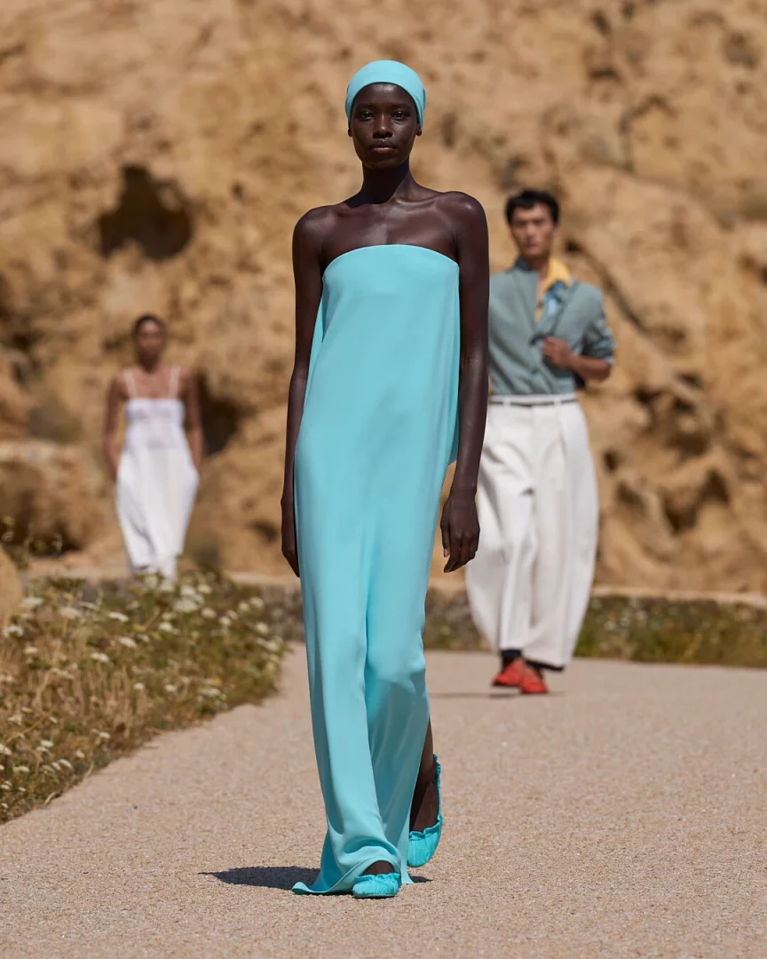 Jacquemus Le Bonheur İlkbahar-Yaz 2027 defilesinden, turkuaz renkte straplez ve yırtmaçlı uzun elbisesi, başörtüsü ve babetleriyle doğal taşlı bir patikada yürüyen bir model, arkasında bulanık iki başka model ve kayalık, açık kahverengi bir fonla, koleksiyonun Akdeniz hafifliğini ve canlı renklerini yansıtan bir görünüm.