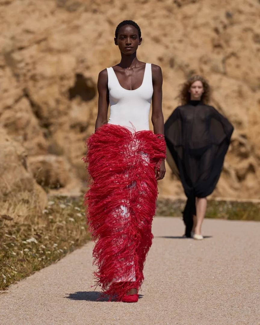 Korsika'nın doğal manzarasında, Jacquemus İlkbahar-Yaz 2027 Le Bonheur koleksiyonundan beyaz askılı bir üst ve rüzgarla dans eden, uçuş uçuş kırmızı püsküllü uzun bir etek giymiş modelin, arkasında bulanık görünen şeffaf siyah bir kıyafetli başka bir modelle yürüdüğü, koleksiyonun hafiflik hissini yansıtan güneşli bir defile anı.