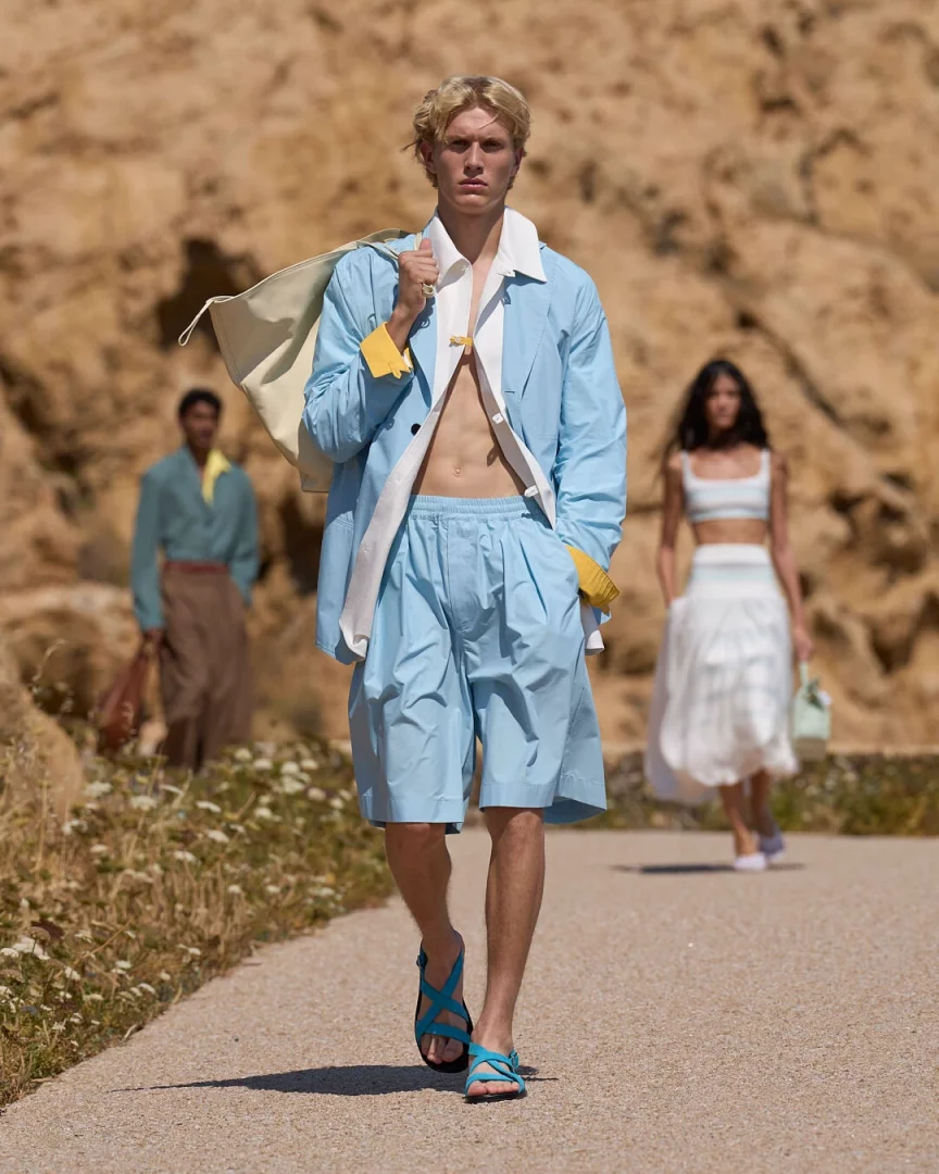 Korsika'nın doğal kayalıkları ve kuru bitki örtüsü arasında, Jacquemus'un Le Bonheur İlkbahar-Yaz 2027 koleksiyonunun hafiflik hissini yansıtan açık mavi bir ceket, pileli şort ve turkuaz sandaletlerle ilerleyen sarışın erkek modelin atletik silueti ve Akdeniz'in canlı renklerinden ilham alan kombini dikkat çekiyor.
