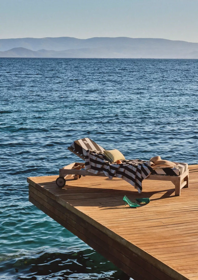 Mandarin Oriental Bodrum'daki Jacquemus beach club iş birliğinin Akdeniz estetiğini sergileyen, siyah beyaz çizgili şezlongun, havlu, hasır şapka ve turkuaz ayakkabıların eşlik ettiği ahşap iskele ve masmavi deniz manzarası.