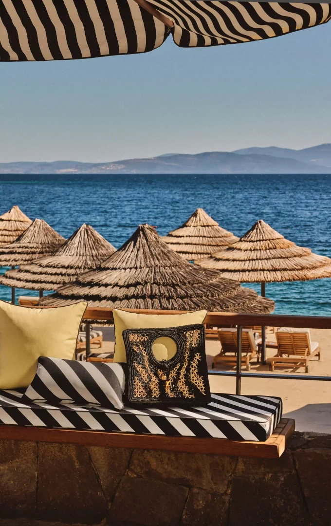 Bodrum'daki Mandarin Oriental Blue Beach Club'ın Jacquemus iş birliği kapsamında, siyah beyaz çizgili şemsiyesi ve minderleriyle dekore edilmiş lüks bir oturma alanında, sarı yastıklar ve üzerinde zarif işlemeli bir çanta görülüyor; arka planda masmavi Ege Denizi ve hasır şemsiyelerle donatılmış kumlu plaj uzanıyor.
