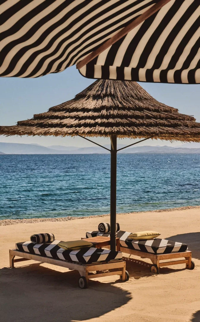 Jacquemus'un 2026 yazı için Mandarin Oriental Bodrum'daki Blue Beach Club iş birliğini yansıtan bu fotoğrafta, siyah beyaz çizgili büyük bir şemsiye, altında saman çatılı bir diğer şemsiye ve üzerinde siyah beyaz zikzak desenli minderleri olan iki ahşap şezlong kumsalın üzerine yerleştirilmiş, arkada masmavi deniz ve dağ manzarası görünüyor.