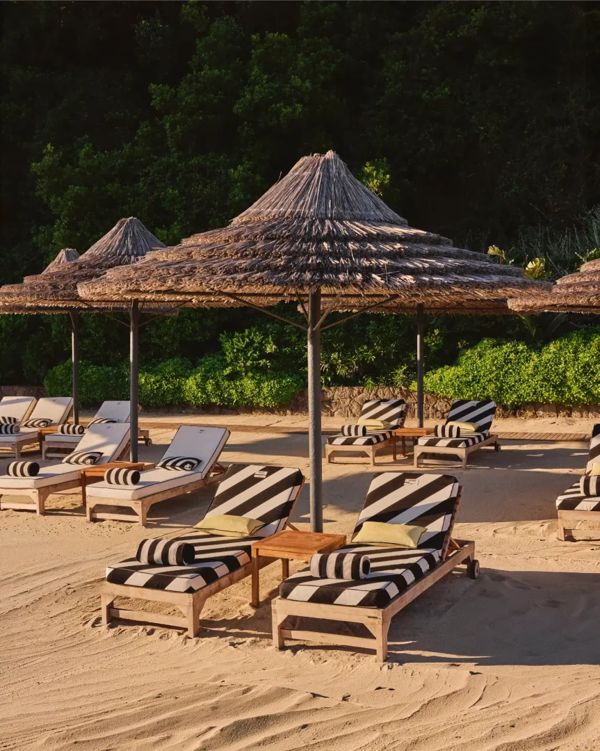 Bodrum'daki Jacquemus x Blue Beach Club iş birliğini yansıtan bu sahil görüntüsünde, kumsalda sıralanmış, markanın çizgili yaz dünyasını temsil eden siyah beyaz desenli şezlonglar ve hasır şemsiyeler güneşin altında tatil atmosferini oluşturuyor.