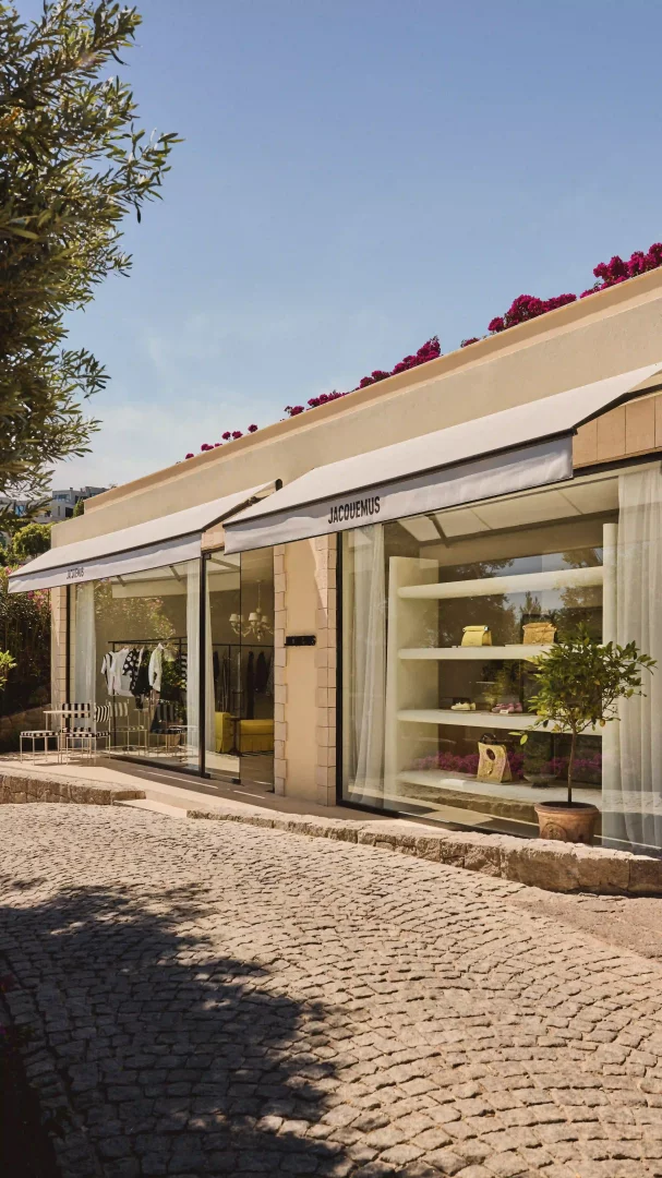 Güneşli bir günde, pembe begonvillerle süslü ve Jacquemus logosunu taşıyan bej cepheli modern bir butiğin vitrininde Resort 2026 Plage koleksiyonundan çantalar ve giysiler sergileniyor; Türkiye'deki ilk moda pop-up mağazası Akdeniz estetiğini Bodrum'a taşıyor.