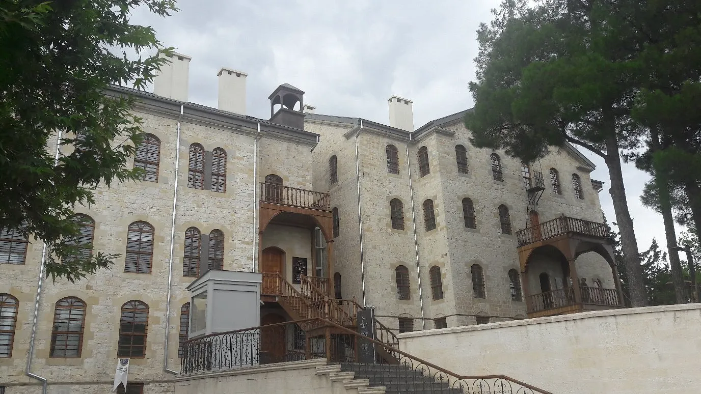 Kahramanmaraş Edebiyat Müzesi'nin geniş, taş duvarlı ana binası, kemerli pencereleri, ahşap balkonları ve ziyaretçi erişimi için eklenmiş modern asansör yapısıyla yeşilliklerin içinde, bulutlu bir gökyüzü altında gösteriliyor.