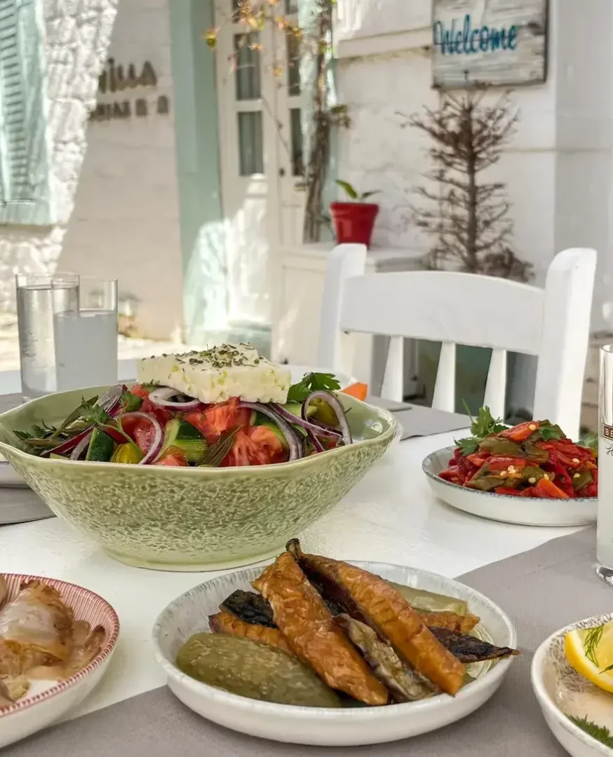 Kızarmış kabak, turşu, közlenmiş kırmızı biber ve küçük balık mezelerinin eşlik ettiği, üzerinde bolca beyaz peynir bulunan iştah açıcı Alaçatı salatası ve rakı kadehleriyle dolu, Ege kasabalarına özgü beyaz badanalı bir binanın önündeki davetkar bir açık hava sofrası.