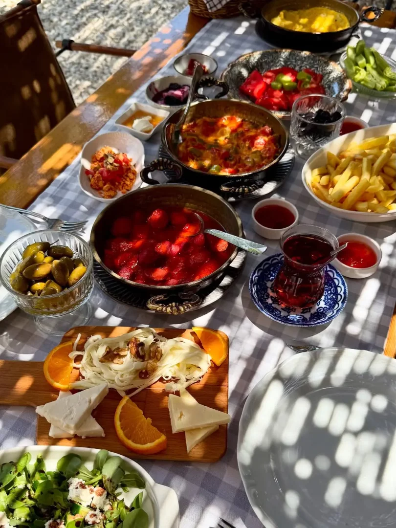 Güneşli bir dış mekanda, ekoseli masa örtülü ahşap bir masada menemen, patates kızartması, çeşitli peynirler, zeytin, çilek reçeli, salata ve demleme çayın yer aldığı, birçok farklı lezzetin bulunduğu bol çeşitli bir kahvaltı sofrası.