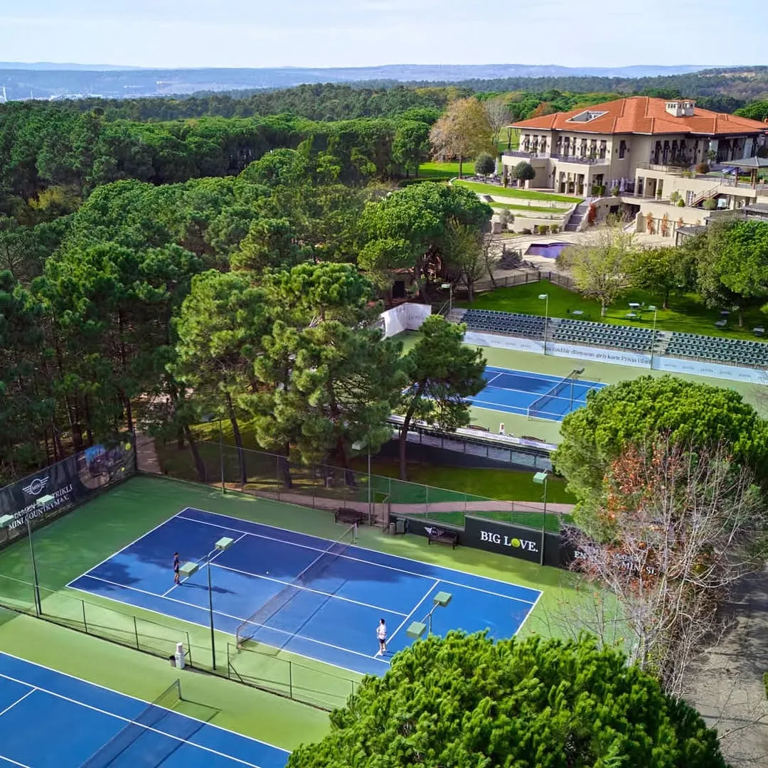 Kemer Country Tenis Kulübü'nün yemyeşil çam ormanlarıyla çevrili kuşbakışı görüntüsünde, birkaç mavi kortta tenis oynayan sporcular, geniş tribünler ve kiremit çatılı lüks bir kulüp binası gün ışığında huzurlu bir ortamda görülüyor.
