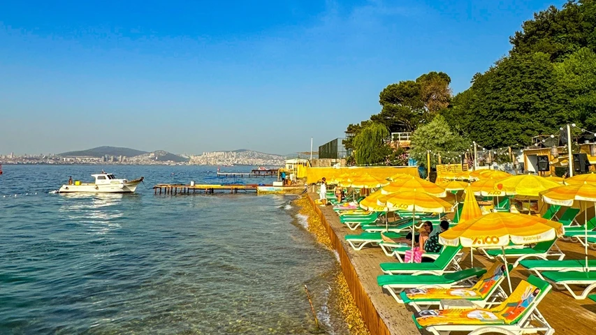 Kınalıada'da denize girilecek yerlerden birini sunan bu parlak yaz gününde, sahil boyunca uzanan ahşap platformda yeşil şezlonglar ve sarı güneş şemsiyeleri sıralanırken, berrak denizde bir tekne yüzüyor ve arka planda ağaçlı tepeler ile şehir manzarası uzanıyor.