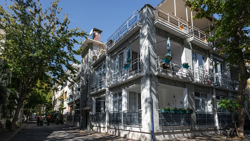 Güneşli bir Kınalıada gününde, balkonlarında mavi saksılar ve küçük şemsiyeler olan, beyaz-gri mozaik kaplamalı çok katlı bir yazlık ev, ağaçların gölgelendirdiği sakin bir sokağın kenarında duruyor ve adanın gezilecek yerlerinden tipik bir manzarayı gözler önüne seriyor.