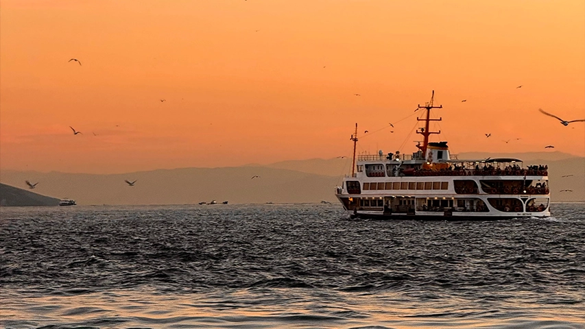 Gün batımının turuncu rengine bürünmüş gökyüzünün altında, Kınalıada gibi adalara ulaşımda sıkça kullanılan, üst güvertesinde ve alt katlarında kalabalık yolcular taşıyan büyük bir vapurun dalgalı denizde ilerlerken martılarla çevrili olduğu görünüyor.