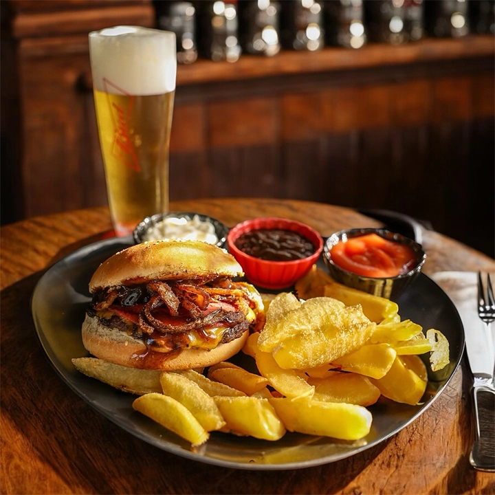 Kınalıada'daki Koala Pub'da bira eşliğinde servis edilen, karamelize soğanlı ve erimiş peynirli bol malzemeli bir burger tabağı, yanında çıtır elma dilim patatesler ve çeşitli soslar ile iştah açıcı bir görüntü sunuyor.