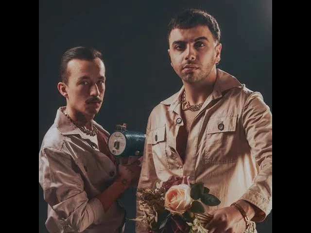 Koyu, sade bir arka plan önünde duran Kofn Grubu üyelerinden, bıyıklı ve saçları geriye taranmış olanı eski model bir fotoğraf makinesi tutarken, diğeri pembe güller ve koyu renkli yapraklardan oluşan bir buketle poz veriyor; ikisi de altın rengi kolyeler takıyor ve bej gömlekler giyiyor.