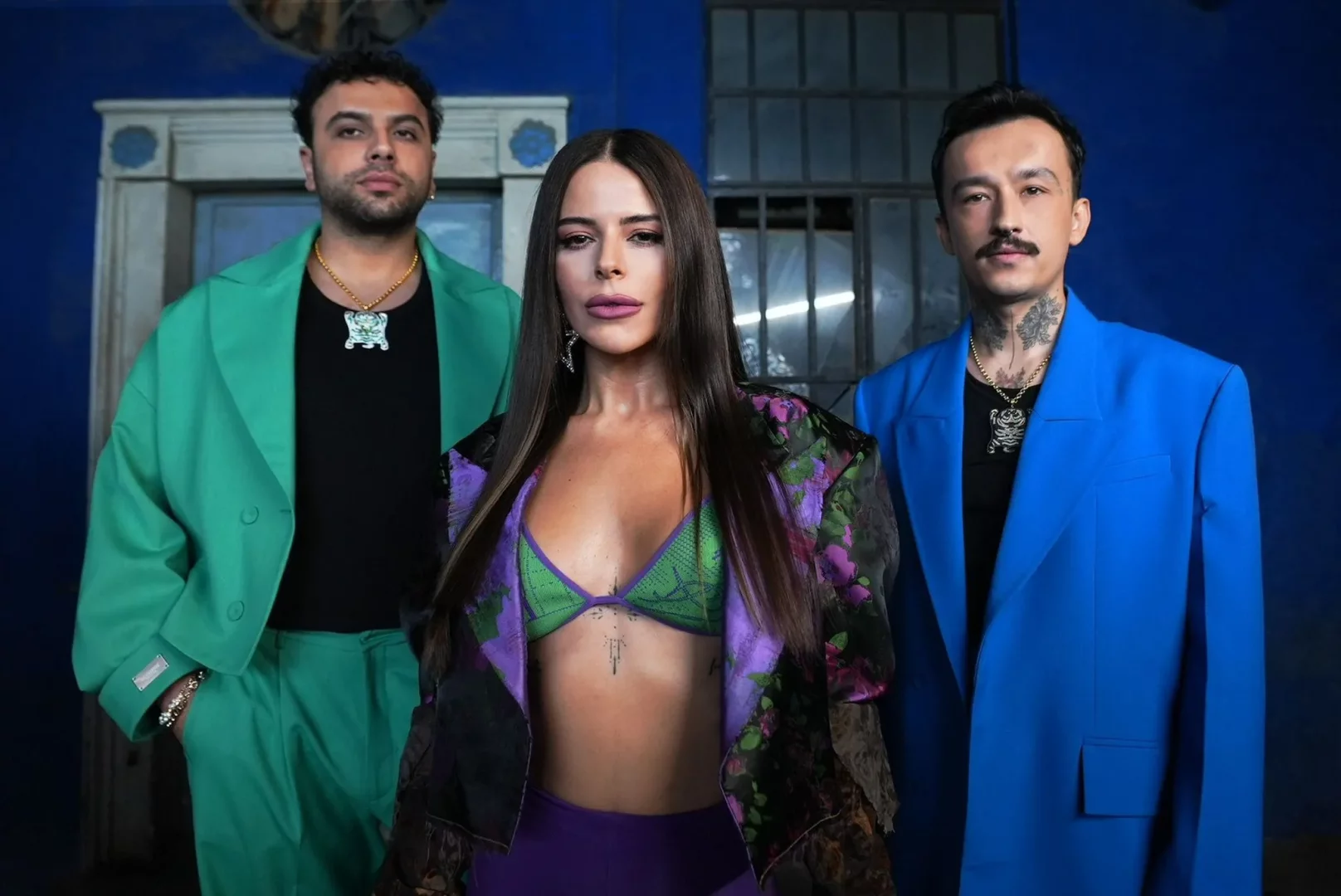 Ortasında canlı yeşil bralet ve mor alt giyen uzun saçlı bir kadının durduğu, sağında dövmeli, bıyıklı bir adamın elektrik mavisi blazer, solunda ise sakallı bir erkeğin parlak yeşil takım elbise giydiği bu karede, üç kişi koyu mavi arka plan önünde çarpıcı ve dikkat çekici tarzlarıyla kararlı bir şekilde objektife bakıyorlar.