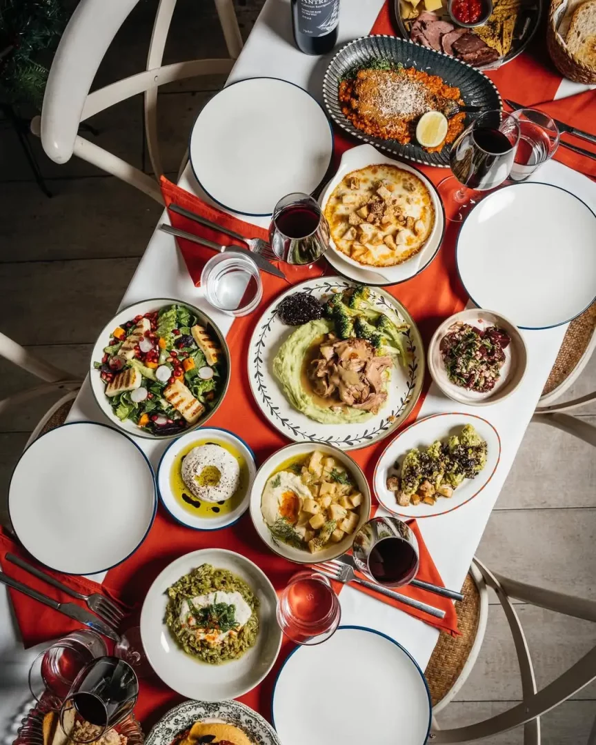 Beyaz masa örtüsü ve kırmızı runner üzerinde, ızgara hellim salatası, etli püre, parmesanlı risotto, çeşit çeşit meze, şarküteri tabağı ve şarap kadehleriyle dolu, davetkar bir ziyafet sofrası yukarıdan göz alıcı bir şekilde sergileniyor.