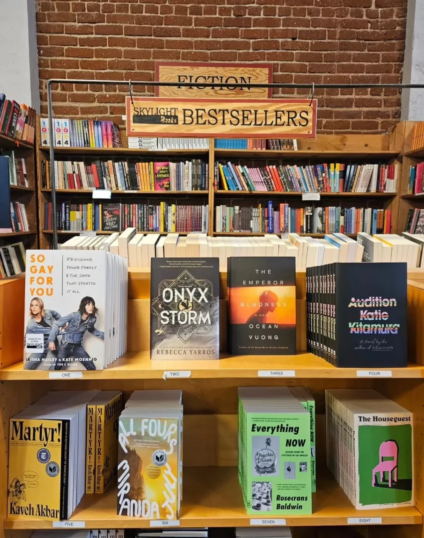 Tuğla duvar fonunda, FICTION ve SKYLIGHT BESTSELLERS yazılı ahşap tabelaların asılı olduğu bir kitapçıda, kapakları öne dönük olarak sergilenen Onyx Storm ve So Gay For You gibi başlıklar dahil olmak üzere, çok sayıda güncel ve çok satan kurgu kitabı dolu iki katlı ahşap raf.