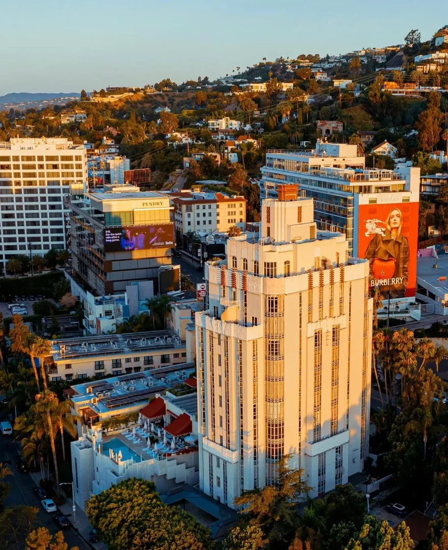 Gün batımının altın rengi ışıklarıyla yıkanan, Art Deco mimarisiyle dikkat çeken, çatısında havuzlu ve kırmızı şemsiyeli bir dinlenme alanı bulunan bu Los Angeles otel binası, palmiye ağaçları ve tepelerle dolu şehrin canlı görüntüsüne hakim.
