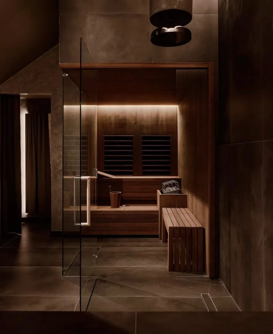 Oteldeki lüks bir dinlenme alanını gösteren bu fotoğrafta, koyu renk ahşap panellerle çevrili, ambiyans aydınlatmalı modern bir sauna odası, içindeki ahşap kova ve kepçe detaylarıyla, ferahlatıcı bir deneyim için misafirlerini bekliyor gibi görünüyor.