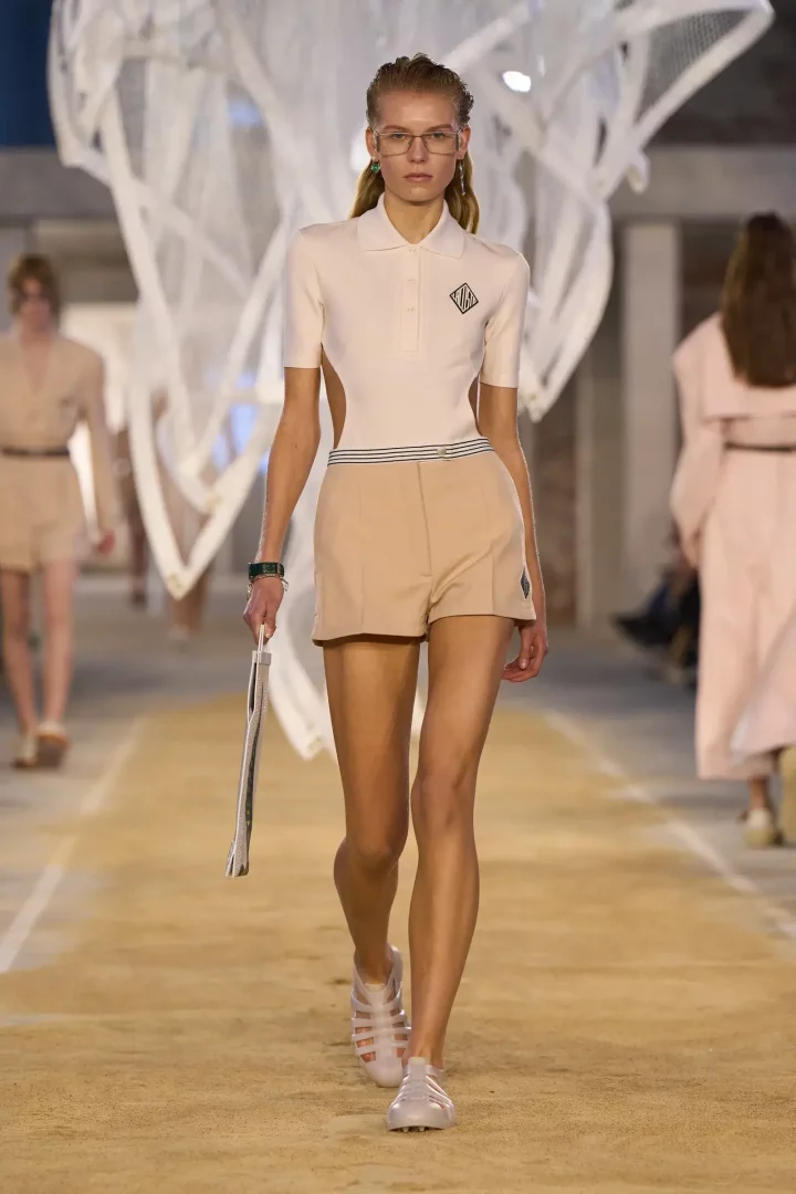 Lacoste İlkbahar 2025 hazır giyim defilesinden bir model, bej yüksek belli şort ve yanları açık krem rengi, logolu bir polo tişört giymiş, şeffaf jöle ayakkabılarıyla podyumda yürüyor, koleksiyonun sportif ama şık ruhunu yansıtıyor.
