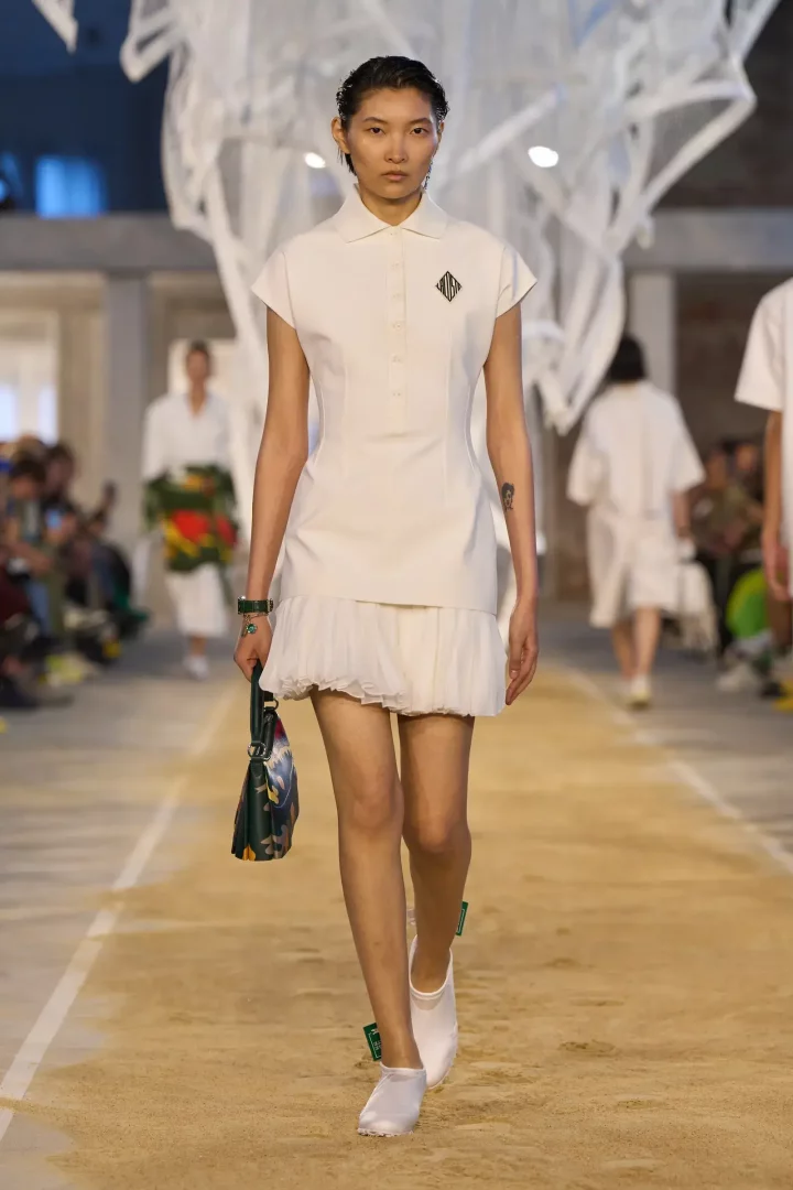 Lacoste İlkbahar 2025 hazır giyim koleksiyonundan bir parçayı tanıtan model, podyumda göğsünde siyah bir logo bulunan, yakalı, kısa kollu, etek ucu büzgülü beyaz mini elbisesi, elinde çiçek desenli koyu yeşil çantası ve beyaz babet benzeri ayakkabılarıyla kumlu zeminde yürürken görülüyor.
