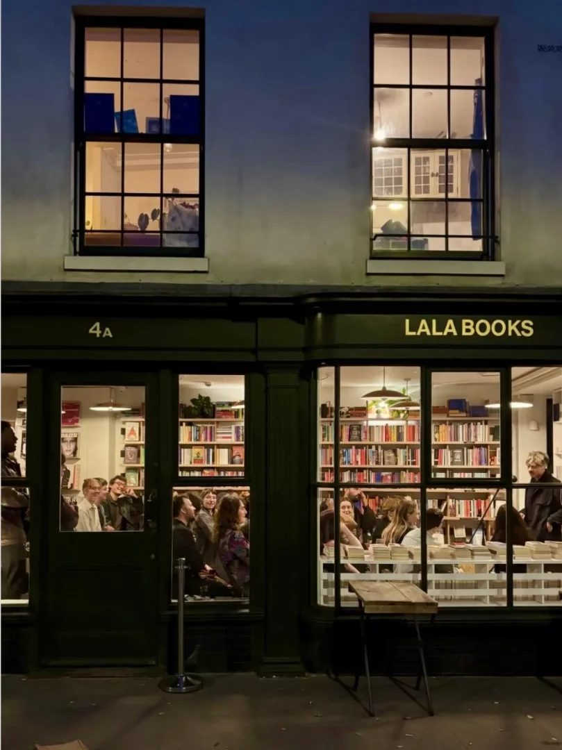 Akşam saatlerinde dışarıdan görünen Lala Books kitabevinin ışıklandırılmış iç mekanında, raflarda renkli kitaplar ve masalarda yeni kitap yığınlarıyla çevrili, neşeli bir kalabalık toplanmış, kültürel bir etkinliğin veya imza gününün canlı atmosferini yansıtıyor.