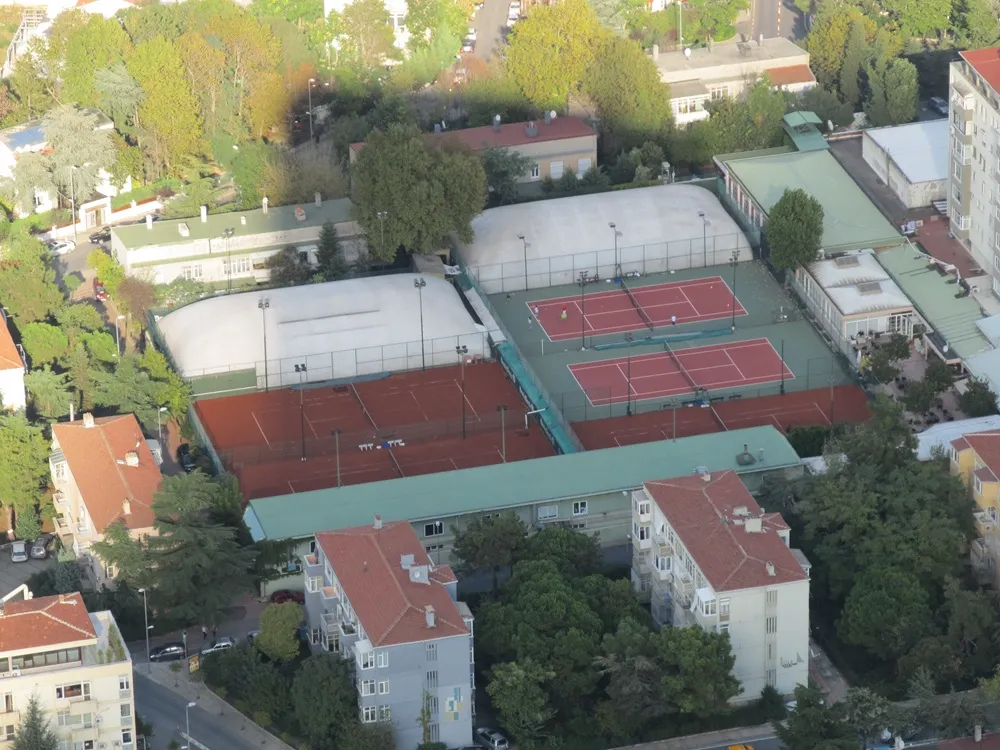 Bir kuşbakışı görüntüde, Levent Tenis Kulübü'nün çevresi ağaçlarla kaplı binalar arasında konumlanmış, üzerinde birkaç kişinin tenis oynadığı iki adet açık kırmızı kort, yanında üç adet toprak kort ve iki adet beyaz kapalı kort olmak üzere toplam yedi tenis kortu içeren geniş alanı görülüyor.