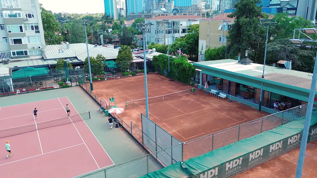 Levent Tenis Kulübü'nün kalabalık şehir manzarasıyla iç içe, yemyeşil ağaçların ve modern binaların çevrelediği, biri pembe diğeri toprak kort olan iki tenis sahasında raket sallayan sporcuları ve sosyal tesislerini gösteren kuşbakışı görünüm.