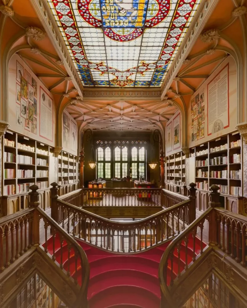 Porto'daki Livraria Lello'nun görkemli iç mekanında, kırmızı halılı zarif kıvrımlı ahşap merdivenler, tavandaki detaylı renkli vitray pencerenin aydınlattığı duvarlarda yükselen, kitaplarla dolu kat kat raflar arasında büyüleyici bir manzara oluşturuyor.