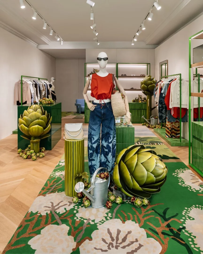 Ortada güneş gözlüklü bir cansız mankenin turuncu bir üst ve palmiye desenli bol paça pantolon giydiği Loewe pop-up mağazasının iç görünümü; yeşil, bitki desenli halı üzerinde dev enginar heykelleri ve sulama kabıyla Paula's Ibiza koleksiyonunun Akdeniz yaşamından ilham alan atmosferini yansıtıyor.