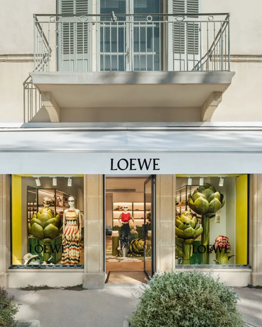 Loewe yazlık koleksiyonunun sergilendiği, ön cephesinde Loewe marka logosu bulunan bir pop-up mağazasının dış görünümü; vitrinlerde devasa enginar heykelleri, güneş gözlüklü mankenler ve renkli çizgili yazlık elbiseler ile Akdeniz yaşamından ilham alan özel bir moda deneyimi sunuyor.