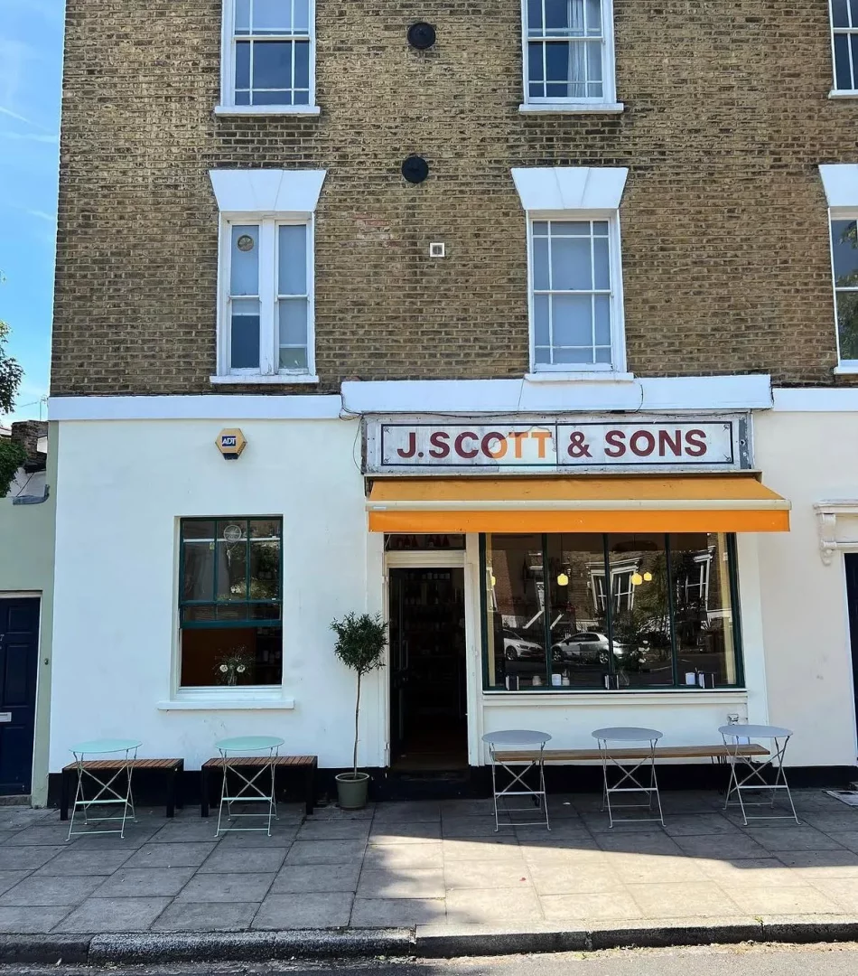 J. SCOTT & SONS adlı Londra barının dış cephesi, tuğla üst katı ve beyaz alt katı ile dikkat çekiyor; büyük vitrin camından yansıyanlar ve portakal rengi tentenin altında, kaldırımda yer alan metal ayaklı masalar, ahşap banklar ve küçük ağaç ile davetkar bir atmosfer sunuyor.