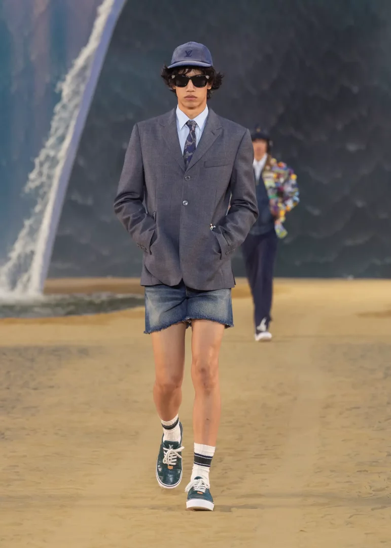 Louis Vuitton İlkbahar-Yaz 2027 Erkek Giyim Defilesi'nde, gri blazer ceket, eskitilmiş denim mini etek, şapka ve spor ayakkabılar giyen bir model, Paris'teki dalga temalı devasa sahne önünde, şehrin ve sahilin kesiştiği modern dandy stilini gözler önüne seriyor.