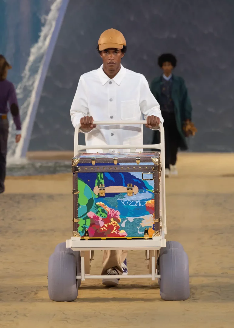 Louis Vuitton İlkbahar-Yaz 2027 erkek giyim defilesinde, Pharrell Williams'ın okyanus ve sörf kültüründen ilham alan koleksiyonundan bir model, kumlu podyumda, canlı mercanlar ve deniz yaşamı desenleriyle süslenmiş lüks bir seyahat sandığını taşıyan beyaz bir el arabasını itiyor.