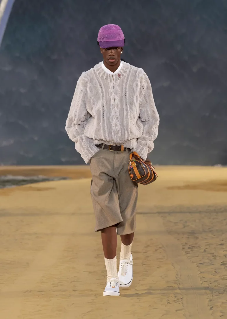 Louis Vuitton 2027 İlkbahar-Yaz Erkek Koleksiyonu'ndan, kum rengi zeminde dalgalı deniz fonunda yürüyen model; mor şapka, açık gri örgü kazak, bej şort ve kemerine asılı küçük Louis Vuitton monogram çanta ile şehirli ve rahat yaz stilini bir araya getiriyor.
