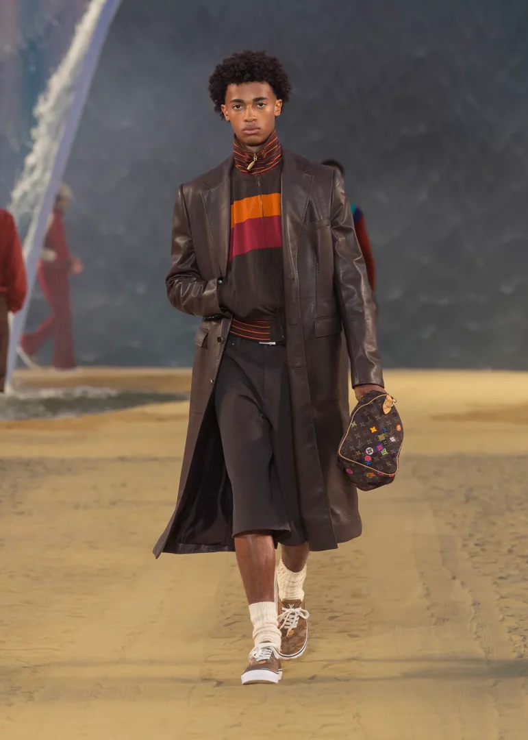 Louis Vuitton İlkbahar-Yaz 2027 erkek koleksiyonundan bir model, dalgalı suların çağrıştırdığı bir defile setinde, uzun kahverengi deri görünümlü bir palto, çizgili fermuarlı bir üst, şort, bilekleri büzgülü çoraplar ve kahverengi spor ayakkabılar giyerken, elinde renkli monogram logolu bir çanta taşıyarak Pharrell Williams'ın hafifleyen terzilik yorumunu sergiliyor.