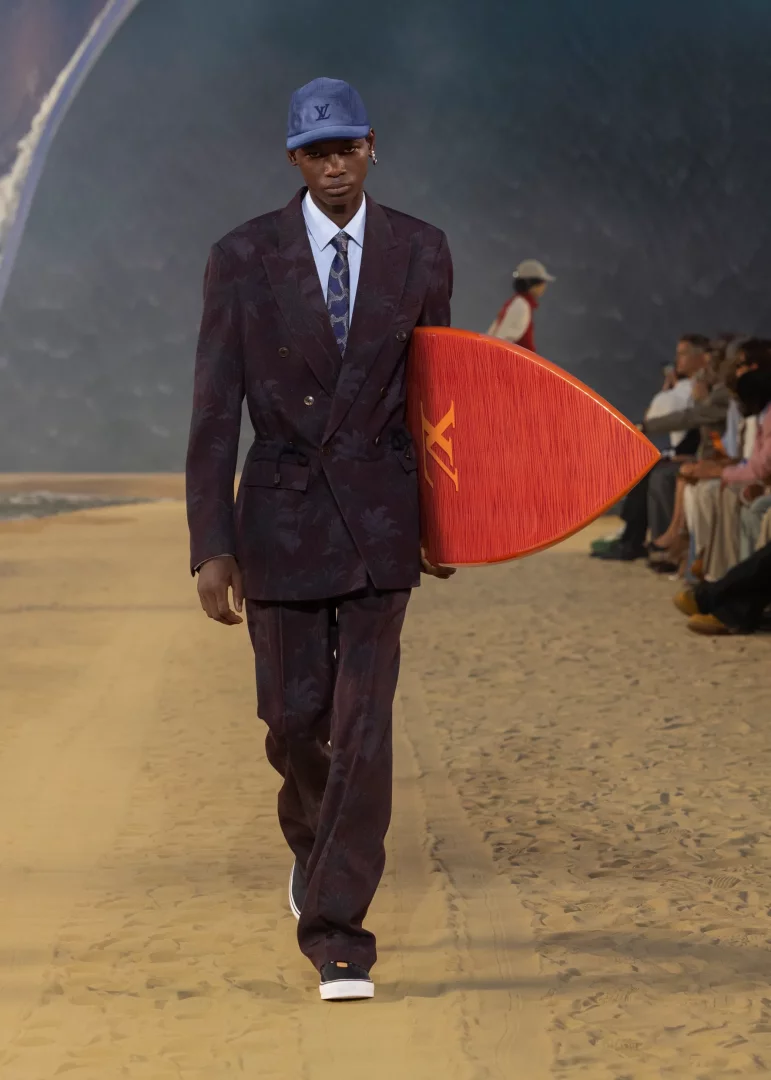 Louis Vuitton 2027 İlkbahar-Yaz Erkek Koleksiyonu defilesinde, kumdan podyumda yürüyen bir model, üzerinde mor yaprak desenleri olan kruvaze bir takım elbise, mavi LV logolu bir şapka ve kravat giyerek, okyanus temalı bir fonda turuncu renkli Louis Vuitton logolu bir sörf tahtası taşıyarak yaz zarafetini ve sahil-şehir stilini birleştiriyor.