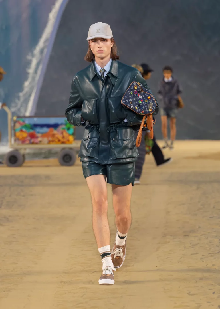 Açık renk şapka, koyu yeşil deri ceket ve şort giymiş, elinde renkli Louis Vuitton monogramlı çanta taşıyan, Paris Moda Haftası'ndaki Louis Vuitton İlkbahar-Yaz 2027 Erkek Giyim Defilesi'nden, hafifleyen terzilik akımını yansıtan bir model podyumda yürüyor.