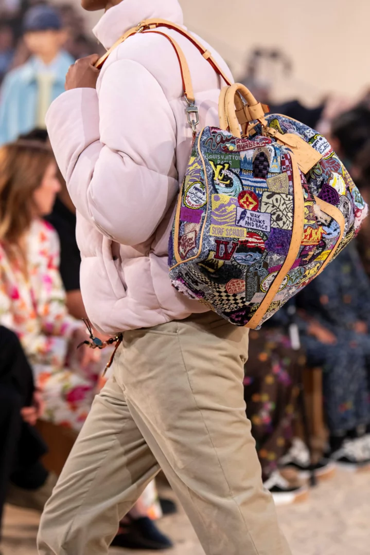 Bir model, Louis Vuitton'un 2027 İlkbahar-Yaz erkek koleksiyonundan sörf kültürü temalı, dalga motifleri ve farklı yamalarla bezeli renkli bir seyahat çantasını omzunda taşırken görülüyor, hafif pembe bir şişme mont ve açık kahverengi pantolonla podyumda yürüyor.