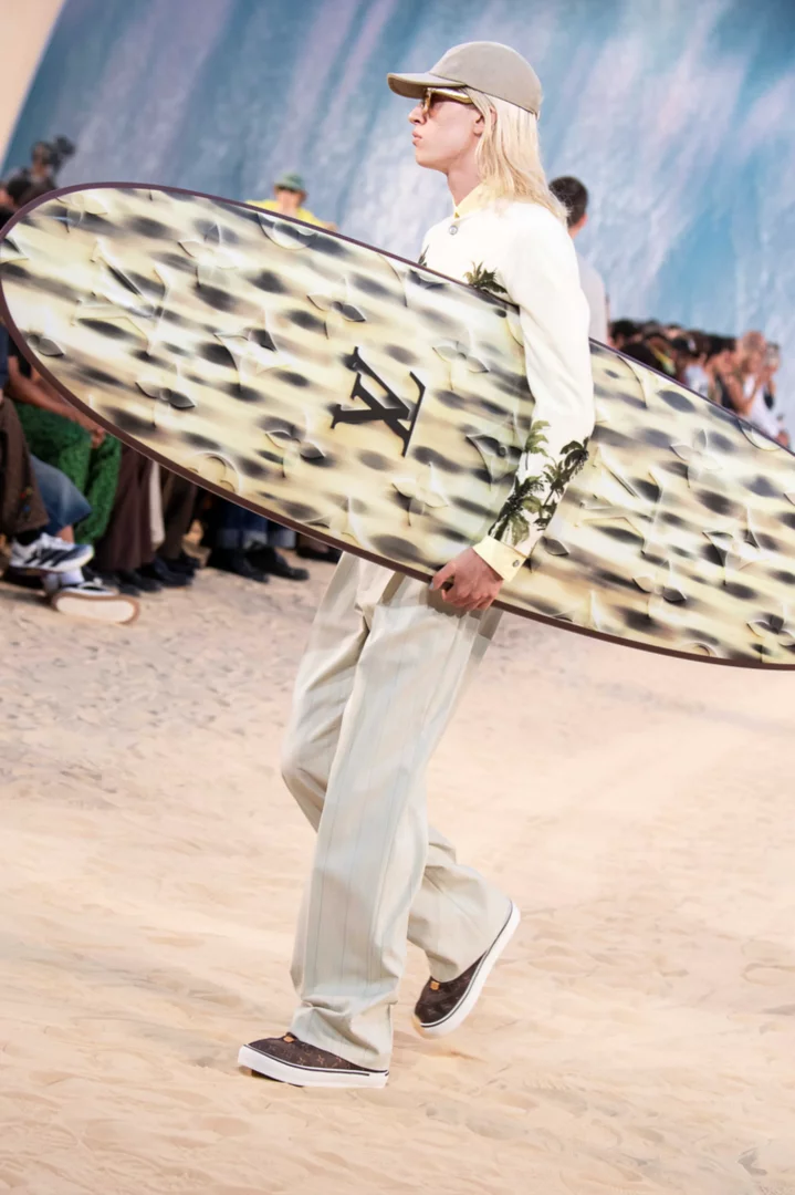 Louis Vuitton 2027 İlkbahar-Yaz Erkek Koleksiyonu defilesinde, kum rengi şapka ve güneş gözlüğü takan, palmiye desenli açık renk bir gömlek, çizgili bol pantolon ve monogram desenli spor ayakkabılar giyen sarı saçlı bir model, üzerinde Louis Vuitton monogramı bulunan sörf tahtasını taşıyarak sahildeki modern dandy imajını yansıtıyor.