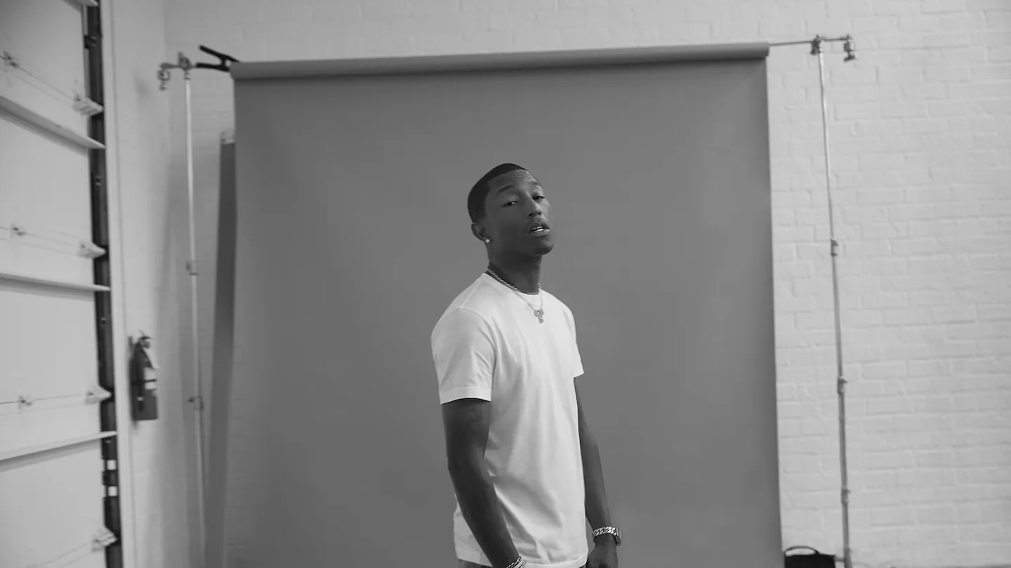 Siyah beyaz bir stüdyo fotoğrafında, beyaz tişört ve dikkat çekici aksesuarlar giyen Pharrell Williams, gri bir fon önünde hafifçe yukarı bakarken Louis Vuitton'un yaratıcı vizyonundan ilham alan duruşuyla objektife poz veriyor.