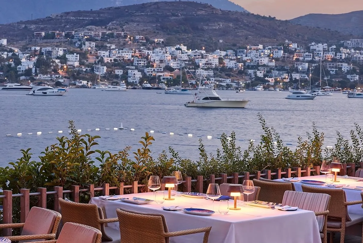 Bodrum'daki Maçakızı'nın akşam alacasındaki teras restoranından çekilen bu görselde, bembeyaz örtülü masalarda şarap kadehleri ve minik masa lambaları eşliğinde zarif bir akşam yemeği atmosferi sergilenirken, arka planda Ege Denizi'nde demirli lüks yatlar ve ışıkları yanan sahil evleriyle çevrili tepeler görünüyor.