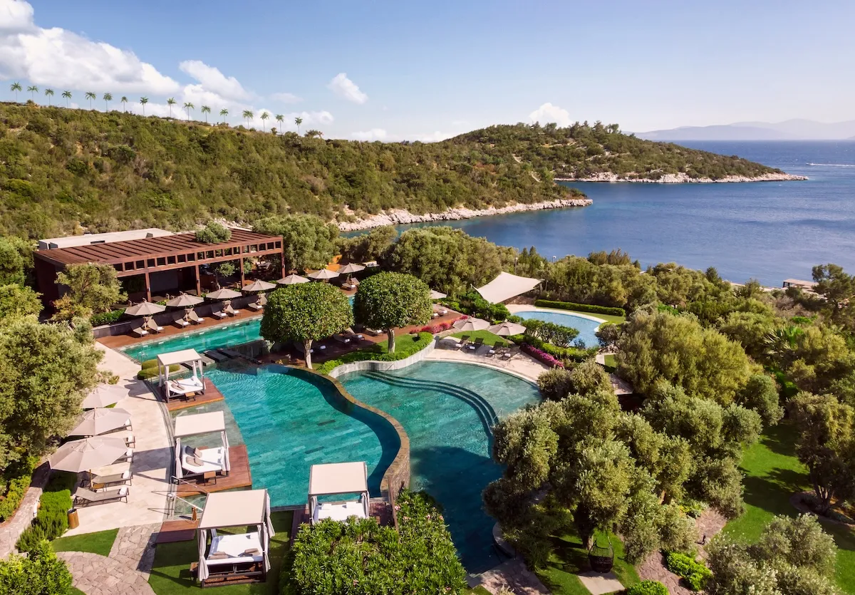 Mavi bir gökyüzü altında, yemyeşil bitki örtüsüyle kaplı tepelerin çevrelediği Cennet Koyu'nun berrak turkuaz sularına bakan Mandarin Oriental Bodrum lüks resort otelini kuşbakışı gösteren bu geniş planda, ahşap teraslar üzerinde şık şezlonglar, gölgelikler, özel dinlenme alanları ve farklı derinliklerdeki kıvrımlı havuzlar net bir şekilde görülüyor.
