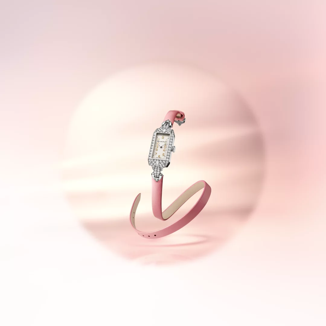 Marilyn Monroe'nun Art Deco etkili orijinal saatinden ilham alan Blancpain Ladybird Tribute koleksiyonuna ait, beyaz altın dikdörtgen kasası pırlantalarla çevrili, opalin kadranlı ve sarı altın indeksli zarif bir kadın saati, şeftali tonunda çift turlu deri kayışıyla yumuşak pembe bir arka plan önünde havada süzülüyor.