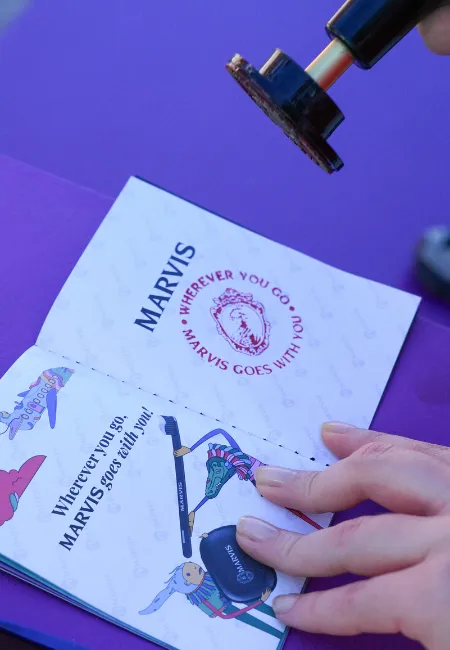 Bir elin, üzerinde Marvis markasının logosu ve WHEREVER YOU GO MARVIS GOES WITH YOU yazısı ile çeşitli seyahat ve ürün illüstrasyonları bulunan bir broşürü tuttuğu görülüyor; mor bir zeminin üzerinde havada duran bir mühür de markaların yaratıcı gelişmelerini ve yeni tasarımlarını gösteriyor.